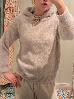 Beige stickad hoodie med snörning - Mysig beige stickad hoodie med huva och snörning i halsen. Tröjan har långärmade och ribbade muddar vid ärmslut och nederkant. Perfekt för dig som gillar en chill och avslappnad stil under kyliga dagar.