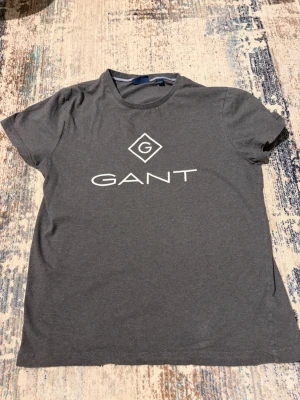 Mörkgrå GANT t-shirt med logga - Säljer en mörkgrå t-shirt från GANT i storlek M. T-shirten har klassisk rund hals och vit GANT-logga tryckt på bröstet. Materialet är mjuk bomull och passformen är normal. Perfekt för dig som gillar stilrena och enkla plagg med tydlig branding.