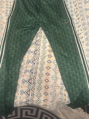 Gröna mönstrade mjukisbyxor Lacoste - Säljer ett par gröna mjukisbyxor från Lacoste med geometriskt mönster och vita ränder längs sidorna. Byxorna har en avslappnad passform och är perfekta för chill dagar. Klassisk Lacoste-logga på benet. Materialet känns mjukt och skönt mot huden.