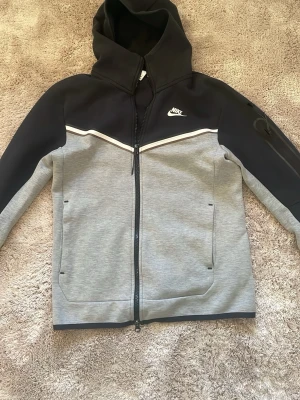 Grå&svart nike tech - Säljer denna nike tech för den är lite liten för mig. Perfekt till dig som vill ha en skön zip hoodie i skolan och fräsch look.