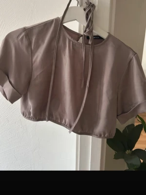 Beige croppad omlottopp Gina Tricot - Säljer en beige croppad topp från Gina Tricot i storlek 34. Toppen har korta ärmar, omlottdetalj med snörning i ryggen och knytband som går runt midjan. Materialet är mjukt och har en lätt glansig yta. Perfekt för dig som gillar trendiga och stilrena plagg.