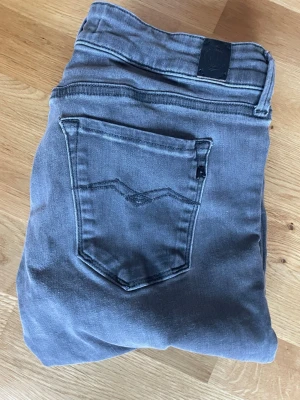 Grå skinny jeans från Replay, storlek 28 32 - Snygga grå jeans från Replay i skinny fit, storlek 28. Klassisk femficksmodell med svarta detaljer och knappstängning. Jeansen har låg midja och är tillverkade i mjukt bomullsmaterial med stretch för extra komfort. Perfekt för dig som gillar en smal siluett.
