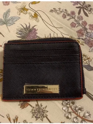 Svart plånbok från Tommy Hilfiger - Snygg svart plånbok från Tommy Hilfiger med guldfärgad logoplatta framtill. Plånboken har flera kortfack och en praktisk dragkedja runt om. Röd kant ger en cool kontrast och insidan har två fack för sedlar eller mynt. Perfekt storlek för fickan!