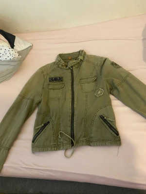 Grön vindjacka med dragkedjor - Snygg grön vindjacka med coola detaljer som snörning i midjan, flera fickor med lock och dragkedjor, samt broderade patches på bröstet och ärmen. Jackan har hög krage med knappar och långärmade ärmar med dragkedjor vid mudden. Perfekt för dig som gillar streetstyle.