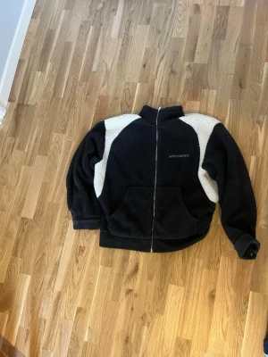 Svart teddyjacka från MADISOn - Svart teddyjacka med vita paneler på axlarna och ärmarna, hel dragkedja framtill och hög krage. Jackan har två stora fickor och ett broderat MADISON-tryck på bröstet. Perfekt för dig som gillar chill och oversized vibe. Säljs inte längre, orginal pris var 1500kr