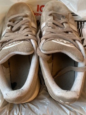 Beige Adidas Campus sneakers - Säljer ett par beige Adidas Campus sneakers med klassiska vita ränder på sidorna och gummisula. Skorna har snörning och är tillverkade i mocka med rund tå. Perfekt för dig som gillar retrostil och vill ha något som funkar till jeans eller shorts.