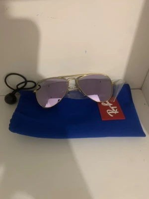Solglasögon för barn ray ban -  ett par Ray-Ban solglasögon med lila glas och guldfärgad metallbåge, samt tillhörande blått fodral och röd Ray-Ban ask.