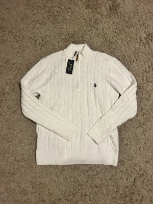 Vit stickad polotröja Polo Ralph Lauren - Vit kabelstickad polotröja från Polo Ralph Lauren med half zip och klassisk broderad logga på bröstet. Tröjan har långärmad design och ribbade muddar. Perfekt för dig som gillar stilrena och tidlösa plagg.