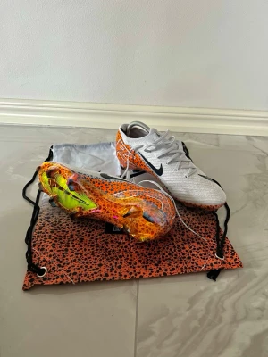 Nike Mercurial Elite 16  - Säljer ett par grymma Nike fotbollsskor i storlek EUR 42. Skorna har en vit ovandel med svarta swoosh-loggor och coolt orange prickigt mönster. Sulan är transparent med neonfärgade detaljer och dobbar. Ovandelen är stickad och har snörning för bästa passform.