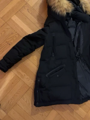 Svart dunjacka med fuskpäls från Rockandble - Svart dunjacka med fluffig fuskpäls på huvan, flera dragkedjefickor och knappar framtill. Jackan har lång ärm och är quiltad för extra värme. Insidan har justerbar midja med snörning. Perfekt för kalla dagar.