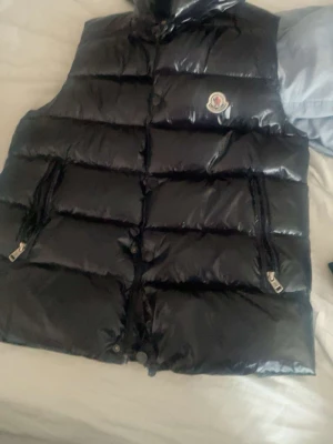 Svart dunväst från Moncler - Säljer en svart, glansig dunväst från Moncler med klassisk logotyp på bröstet. Västen har quiltad design, två fickor med dragkedja och hög krage. Perfekt för lager-på-lager och har en riktigt clean look som funkar till jeans och hoodie.