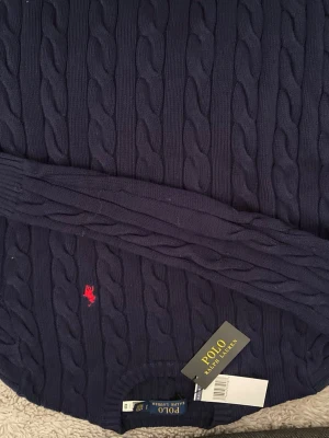Marinblå kabelstickad tröja Polo Ralph Lauren - Säljer en marinblå kabelstickad tröja från Polo Ralph Lauren i 100% bomull. Tröjan har långärmad design, ribbade muddar och rund hals. Klassisk röd broderad logga på bröstet. Perfekt för dig som gillar stilrena och tidlösa plagg.