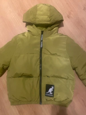 Olivgrön pufferjacka från Kangol limited edition - Säljer en olivgrön pufferjacka från Kangol med huva och svart logotyp på bröstet. Jackan har en rak passform, hel dragkedja framtill och elastisk nederkant. Materialet är syntet och jackan är perfekt för kyliga dagar. Finns inte längre att köpa då den var limited edition