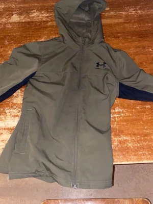 Grön vindjacka från Under Armour - Säljer en grön vindjacka från Under Armour med svarta detaljer längs ärmarna. Jackan har huva, dragkedja framtill och två sidofickor. Perfekt för blåsiga dagar och har en sportig look med loggan på bröstet.