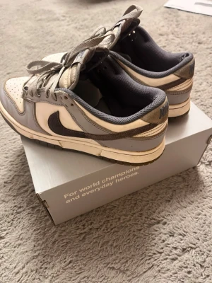 Nike Dunk Low i grå och vitt - Nike Dunk Low sneakers i en clean kombo av grått och vitt. Ovandelen är i skinn med grå mocka-swoosh och matchande grå snörning. Låg siluett och klassisk perforerad tåbox för extra vibe. Insidan är fodrad i mörkgrått tyg. Perfekt för dig som gillar retrostil.