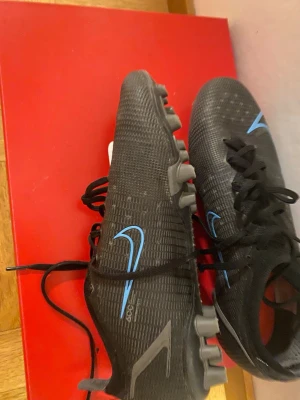 Nike svarta fotbollsskor med blå detaljer - Säljer ett par svarta Nike fotbollsskor med blå swoosh och snörning. Skorna har inte använts mycket och  är exklusiva och går inte att köpa pris kan diskuteras 