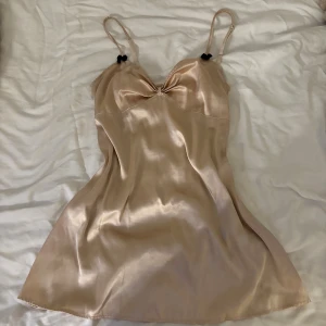 Champagnefärgad satin nattlinne med rosetter - Säljer ett glansigt champagnefärgad nattlinne i satin med tunna axelband och små svarta rosetter vid banden. Super mjuk och fin🩷Passar mig som har xs/s👌🏻