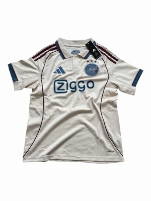 Vit Ajax fotbollströja  - Ajax fotbollströja från Adidas i vitt med vinröda och blå detaljer. Tröjan har klassisk krage, broderat Ajax-emblem och Ziggo-logga på bröstet. Tillverkad i lätt polyester med AEROREADY-teknologi för bästa komfort på planen.