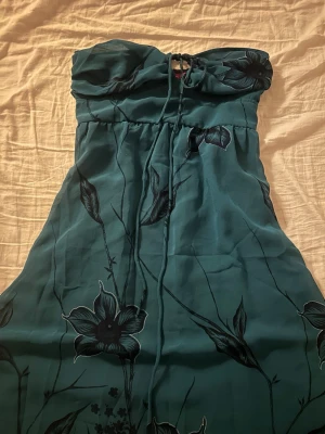 Grön långklänning med snörning - Säljer en grön bandeau långklänning från monsoon med stora blommor i svart och grönt. Klänningen har snörning framtill vid bysten och dragkedja i sidan för enkel påklädning. Tyget är lätt och flowy, perfekt för varma dagar.