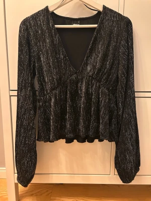 Glittrig svart blus med v-ringning - Supersnygg svart blus från Gina Tricot med glittriga silvertrådar. Djup v-ringning, långa ärmar med en form av ballong ärmar. Perfekt för att sticka ut med en festlig look. Materialet är mjukt och stretchigt, vilket gör den bekväm att bära hela kvällen. Toppen är i storlek S🩷