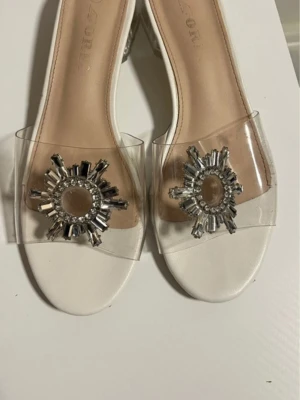 Vita sandaler med strassdetalj - Säljer ett par vita sandaler med transparent rem och stor glittrig strassdekoration framtill. Skorna har öppen tå och en sula med liten klack, vilket ger en elegant och trendig look. Perfekta för dig som vill sticka ut med lite extra bling.