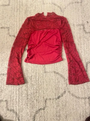 Röd spetsblus med lång ärm - Säljer en röd blus med genombruten spets och långärmad design. Toppen har en öppen urringning. Spetsen har ett blommigt mönster och tyget under är slätt och stretchigt. Perfekt för dig som vill sticka ut lite extra.