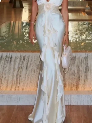 Beige silkes  balklänning med volanger  - Elegant vit beige långklänning i glansigt material med djup v-ringning och volangdetaljer längs hela framsidan. Klänningen har markerad midja och en dekorativ blomma framtill. Perfekt för dig som vill sticka ut på bal. Hel slutsåld !!! Köpt för 1500kr 🥰 fårn Bella barnett