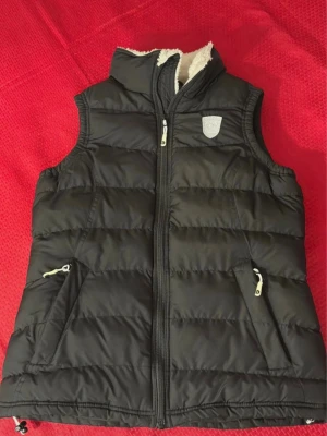 Svart dunväst från Dobsom, strl 36 - Säljer en svart dunväst från Dobsom med mysigt vitt foder, hög krage och två fickor med dragkedja. Västen har en snygg quiltad design och logga på bröstet. Perfekt för lager-på-lager under kyliga dagar.