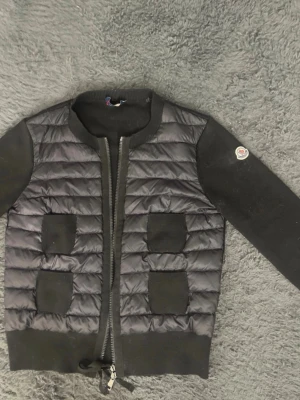 Svart quiltad pufferjacka Moncler - Säljer en svart pufferjacka från Moncler i storlek S. Jackan har quiltad front i blankt material, stickade ärmar och ribbade muddar. Fyra fickor framtill och dragkedja hela vägen. Klassisk Moncler-logga på ena ärmen. Har ett litet hål på baksidan vid nacken men inget som syns. Skriv gärna dm ifall du skulle vilja ha mer bilder på cardiganen eller frågor.