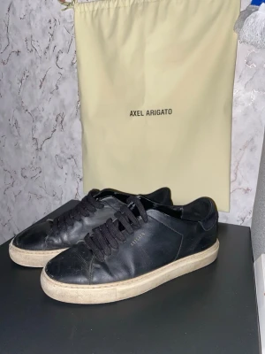 Svarta sneakers från Axel Arigato - Säljer ett par klassiska svarta sneakers från Axel Arigato med vit sula och Axel Arigato gulddetalj på sidan. Skorna har snörning och är tillverkade i skinn, vilket ger en clean och stilren look. Perfekta till jeans eller linnebyxor. Lite använda, men i bra skick för att användas. Tvättar också skorna innan jag skickar dem.(kanske skulle vart bra o göra innan man la ut men)….! Dem andra skorna i bakgrunden på sista bilden är också till salu för rätt pris. De bara o höra av sig vid frågor!