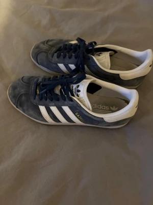 Adidas Gazelle blå i mocka - Säljer ett par klassiska Adidas Gazelle sneakers i blå mocka med vita detaljer. Skorna har snörning, vit sula och guldfärgad Gazelle-text på sidan || Pris: 499|| Storlek: 43 1/3 ||