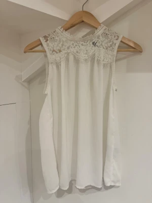 Vit blus med spetsdetaljer - Supersöt vit blus med ärmlös design och transparent spets upptill. Blusen har knäppning i nacken och ett flowy fall. Perfekt för dig som gillar romantiska detaljer och vill ha något luftigt till sommaren.