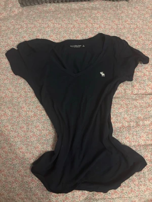 Svart v-ringad t-shirt Abercrombie - Svart t-shirt från Abercrombie & Fitch med v-ringning och liten vit logga på bröstet. Klassisk passform och mjukt bomullsmaterial som känns skönt mot huden. Perfekt till jeans eller shorts för en enkel och clean look.