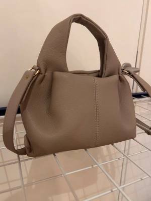 Beige axelväska i skinnimitation - Säljer en stilren beige axelväska i skinnimitation med justerbar axelrem och korta handtag. Väskan har enkel design med guldfärgade metalldetaljer och synlig söm mitt på framsidan. Perfekt storlek för det viktigaste och passar till många outfits.