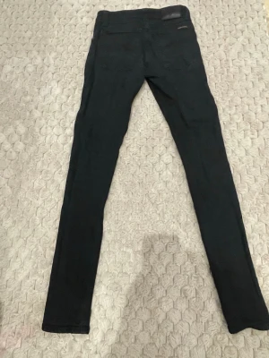 Svarta skinny jeans Nudie Jeansco W26 L32 - Säljer ett par svarta skinny jeans från Nudie Jeans co i storlek W26 L32. Jeansen har klassisk femficksdesign, silverfärgade nitar och knapp, samt smal passform hela vägen ner. Materialet är stretchigt jeans för en bekväm känsla.