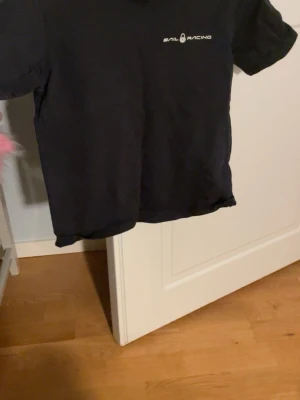 Svart t-shirt från Sail Racing - En svart t-shirt från Sail Racing med liten vit logga på bröstet. Klassisk passform och rund halsringning. Perfekt till jeans eller shorts för en clean look.