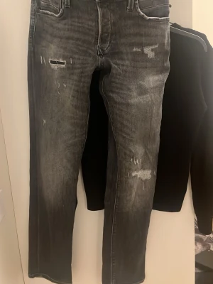 Jack&jones  - Säljer ett par grå jeans med slitningar och lagade detaljer på låret och knät. Jeansen har raka ben och klassisk femficksdesign. Perfekta för dig som gillar en lite ruffare streetstil.