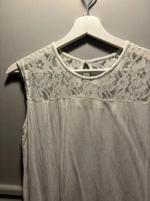 Vit ärmlös topp med spetsdetalj - Säljer en vit ärmlös topp med rund halsringning och vacker spets upptill. Spetsen har ett blommigt mönster som ger en romantisk känsla. Toppen är lätt och luftig, perfekt för varma dagar eller lager på lager. Knäppning bak i nacken för extra detalj.