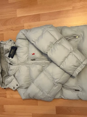 Ljusgrå pufferjacka från Polo Ralph Lauren - Säljer en ljusgrå pufferjacka från Polo Ralph Lauren med huva och flera dragkedjeförsedda fickor. Jackan har det klassiska röda Polo-logot på bröstet och quiltad design. Perfekt för kalla dagar och riktigt snygg till streetwear.