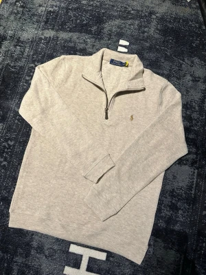 Beige half zip tröja Polo Ralph Lauren - Säljer en stilren beige tröja från Polo Ralph Lauren med klassisk broderad logga på bröstet. Tröjan har half zip-dragkedja, hög krage och långa ärmar. Perfekt för dig som gillar en clean och tidlös look.