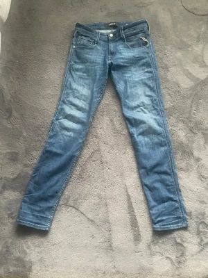 Replay jeans - Säljer ett par blå jeans från Replay med klassisk femficksdesign och snygga slitningar. Jeansen har en mörkblå tvätt, silverfärgad Replay-detalj på bakfickan och ett stort läderpatch bak i midjan. Tillverkade i mjukt och stretchigt jeansmaterial. Hör gärna av dig vid frågor!
