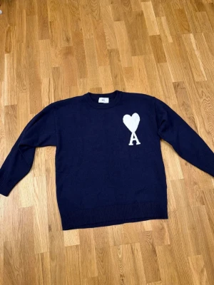Mörkblå stickad tröja från Ami  - Säljer en mörkblå stickad tröja från Ami Alexandre Mattiussi med ikoniskt hjärta och A på bröstet. Tröjan har rund hals, ribbade muddar och är tillverkad i en mjuk ullblandning. Perfekt för dig som gillar stilrena detaljer och vill ha något klassiskt i garderoben.