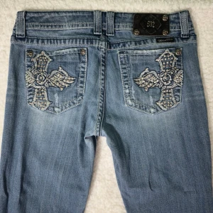 Miss Me ljusblå bootcut jeans - strlk 31 - Säljer ett par ljusblå jeans från Miss Me med bootcut passform. Jeansen har slitningar framtill och stora broderade kors med strassdetaljer på bakfickorna. Klassisk femficksmodell med dekorativa nitar och kontrastsömmar. Perfekta för dig som gillar statement-plagg.