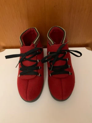Röda norska kängor i ull, storlek 38 - Unika röda kängor från Norway i ull med svart sula och svarta snören. Dekorativ kant i rött, grönt, vitt och gult ger en traditionell vibe. Grov sula för bra grepp och metallhakar för snörningen. Perfekta för dig som vill sticka ut med klassisk nordisk stil.