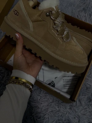 Beige sneakers från UGG med snörning - Säljer ett par chunky beige sneakers från UGG med grov sula och fluffigt foder. Skorna har detaljer i mocka och mesh, samt breda snören i beige och vitt mönster. Perfekta för dig som vill ha både stil och komfort i höst och vinter.