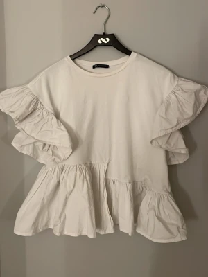 Vit volangblus från Zara - Supersöt vit blus från Zara med stora volangärmar och lös passform. Tillverkad i mjuk bomull och har rund halsringning. Perfekt för dig som gillar en luftig och trendig stil med extra volym i ärmarna. Finns lite sminkfläckar på tröjan men går bort i tvätten!!