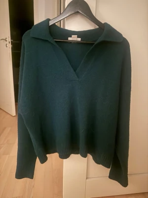 Mörkgrön stickad tröja från Monki - Mysig mörkgrön stickad tröja från Monki i storlek M. Tröjan har en avslappnad krage, v-ringning och långa ärmar med ribbade muddar. Perfekt för lager på lager under kyliga dagar.