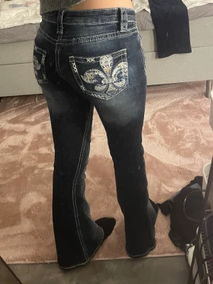 Mörkblå bootcut jeans  - Säljer ett par mörkblå jeans med bootcut passform och snygga broderade fickor bak. Jeansen har kontrastsömmar och låg midja, samt coola detaljer med broderat mönster i vitt och beige på bakfickorna. Perfekta för dig som vill sticka ut lite extra!