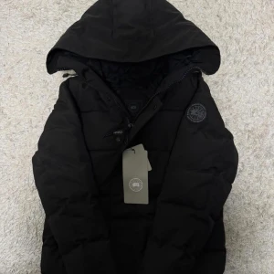 Svart pufferjacka från Canada Goose - Säljer en svart pufferjacka från Canada Goose med huva och diskret logotyp på ärmen. Jackan har dragkedja och knappar framtill samt en quiltad design som håller värmen. Perfekt för kalla dagar och riktigt snygg till alla outfits.