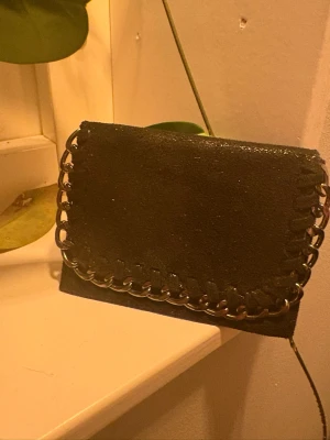 Svart plånbok med kedjedetalj - Säljer en svart plånbok i mocka med glansig finish och silverfärgad kedja runt kanterna. Plånboken har flera kortfack och stängs med tryckknapp. Perfekt storlek för att ha med sig i fickan eller väskan.